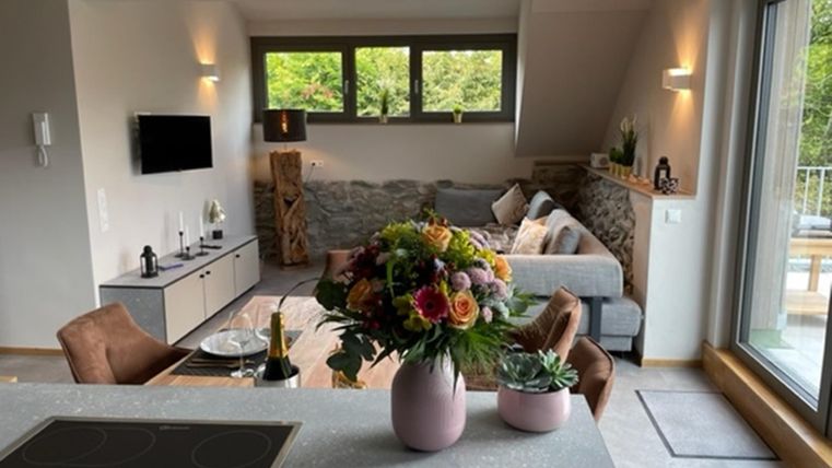 Ein modernes Wohnzimmer mit einem gemütlichen Sofa und großen Fenstern, die viel Licht hereinlassen. Ein schöner Blumenstrauß in einer Vase steht auf dem Tisch.