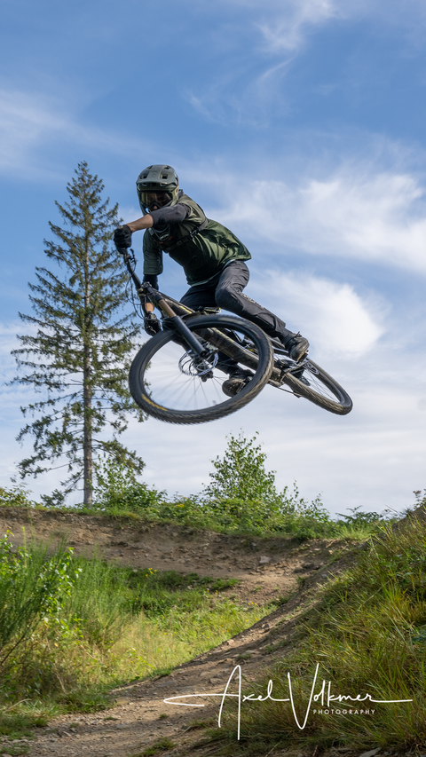 Ein Mountainbiker springt mit seinem Rad über einen Hügel. Im Hintergrund sind Bäume und ein blauer Himmel zu sehen.