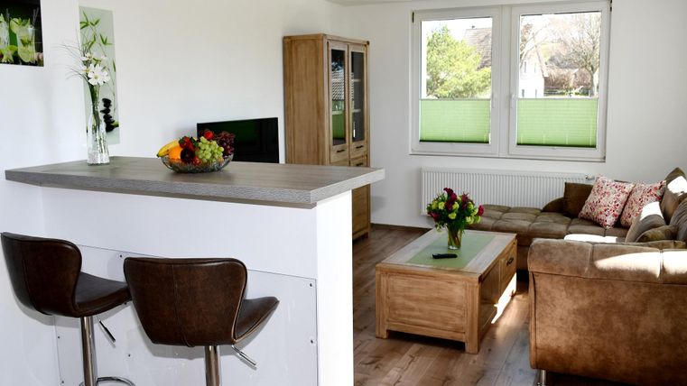 Eine helle und moderne Wohnküche mit einem gemütlichen Sofa und einem Couchtisch. Im Hintergrund sieht man große Fenster, die viel Tageslicht hereinlassen.