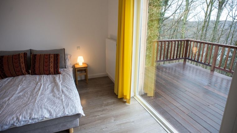 Ein gemütliches Schlafzimmer mit einem Bett und farbenfrohen Kissen. Große Fenster bieten einen Blick auf den Balkon und die Bäume draußen.