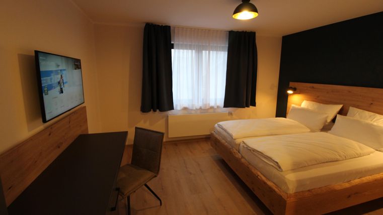 Ein modernes Schlafzimmer mit zwei Betten und einem Schreibtisch. Zum Raum gehört ein Fernseher und große Fenster mit Vorhängen.