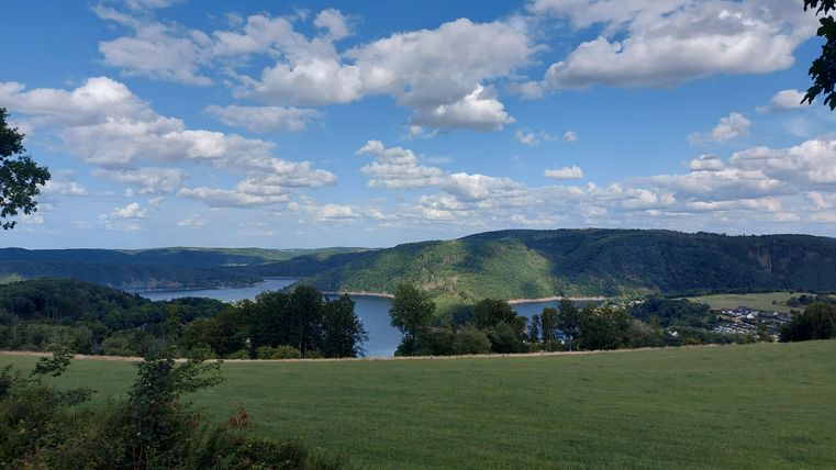 Eine malerische Landschaft mit einem ruhigen See und sanften Hügeln. Der Himmel ist blau und mit vielen Wolken geschmückt.
