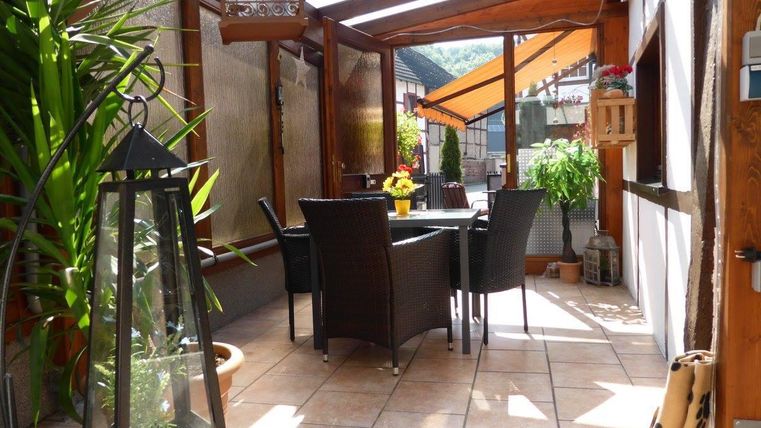 Ein gemütlicher Terrassenbereich mit einem Tisch und Stühlen aus Rattan. Die Umgebung ist grün mit Pflanzen und einer großen Laterne.