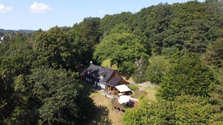 Ein schönes Haus umgeben von grünen Bäumen. Im Vordergrund ist eine Terrasse mit einem Sonnenschirm zu sehen.