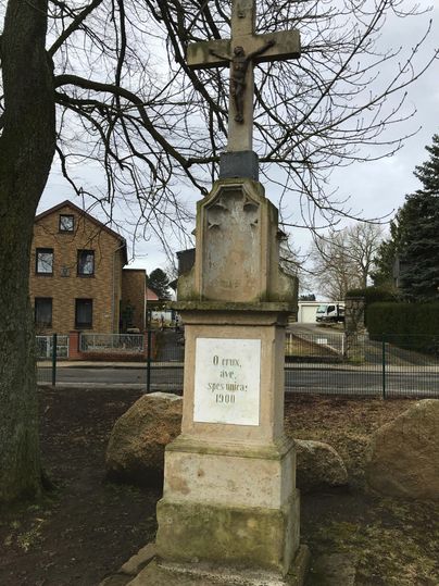 Ein historisches Kreuz steht auf einem Sockel aus Stein. Im Hintergrund sind einige Gebäude und Bäume sichtbar.