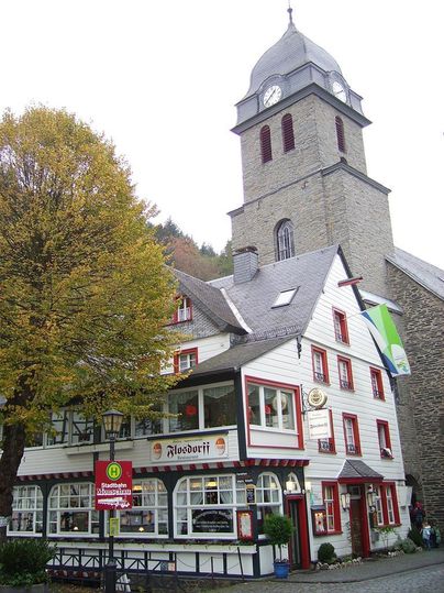 Ein charmantes, zweigeschossiges Gebäude mit einem Uhrenturm im Hintergrund. Bunte Fenster und eine herbstliche Baumkulisse verleihen der Szene eine gemütliche Atmosphäre.
