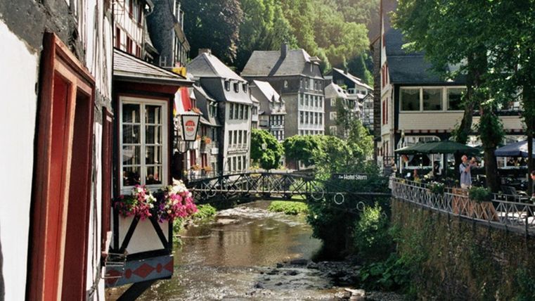 Eine malerische Stadt am Fluss mit traditionellen Häusern und blühenden Blumen. Die umliegende Natur ist grün und einladend.