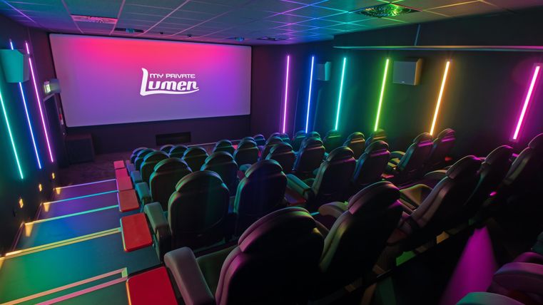 Ein modernes Kino mit komfortablen Sitzplätzen und farbenfroher Beleuchtung. Die Leinwand zeigt das Logo „Lumen“ in einem stilvollen Ambiente.