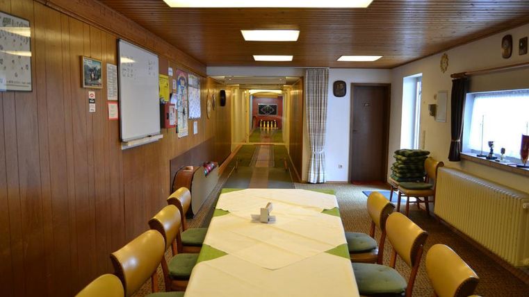 Ein langer Flur mit Holzwänden, der zu einem hellen Raum führt. In der Mitte steht ein gedeckter Tisch mit Stühlen.