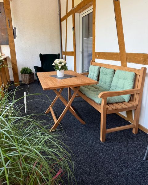 Eine gemütliche Sitzecke auf einem Balkon mit einem Holztisch und einer Bank. Auf dem Tisch steht eine kleine Blumenvase und im Hintergrund sind Pflanzen sichtbar.