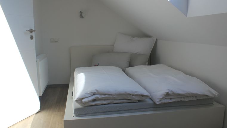 Ein helles Schlafzimmer mit einem weißen Bett und zwei Kissen. Der Raum hat eine gemütliche Atmosphäre und Dachfenster, die Tageslicht hereinlassen.