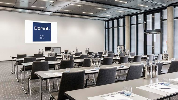 Ein modernes Konferenzzimmer mit Tischen und Stühlen, bereit für ein Meeting. An der Wand hängt ein Bildschirm mit dem Logo des Dorint Hotels.