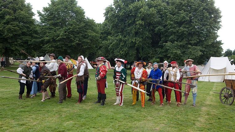 Eine Gruppe von Menschen in historischen Kostümen steht im Freien. Sie halten lange Stäbe und stehen vor Zelten in einem grünen Feld.