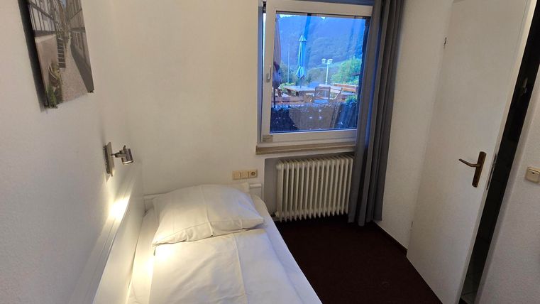 Ein einfaches Einzelzimmer mit einem Bett und einem Fenster. Die Wände sind hell, und es gibt einen Heizkörper sowie einen Schrank.