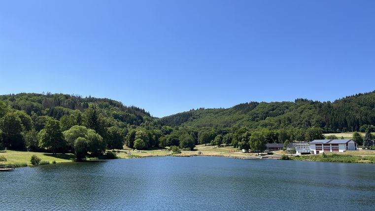 Ein ruhiger See umgeben von grünen Hügeln und Bäumen. Der Himmel ist klar und blau.