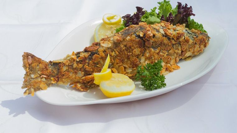 Ein gebratener Fisch auf einem weißen Teller, garniert mit Zitronenscheiben und frischem Salat. Die knusprige Kruste ist mit Cornflakes dekoriert.