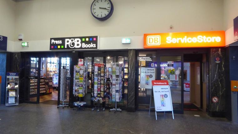Ein Bereich in einem Bahnhof mit zwei Geschäften, einem Buchladen und einem Service-Store. An der Wand hängt eine große Uhr.