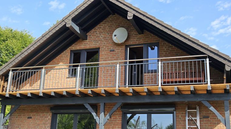 Ein modernes Haus mit einer Holzveranda und großen Fenstern. Auf dem Balkon stehen sichtbare Geländer und eine Sat-Anlage ist installiert.