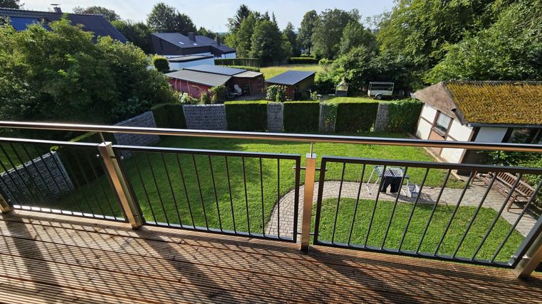 Ein Blick von einem Balkon auf einen schönen Garten mit grünem Rasen und einigen Bäumen. Im Hintergrund sind Häuser und ein gepflasterter Bereich zu sehen.