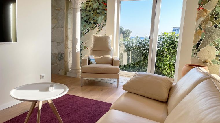 Ein modernes Wohnzimmer mit einem beigen Sofa und einem kleinen weißen Tisch. Große Fenster bieten einen Blick auf die grüne Umgebung und lassen viel Tageslicht herein.