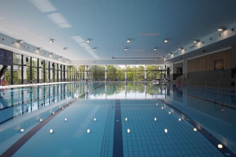 Ein modernes Hallenbad mit einem klaren Wasserbecken. Große Fenster lassen viel Tageslicht herein und bieten einen Blick auf die Außenanlage.