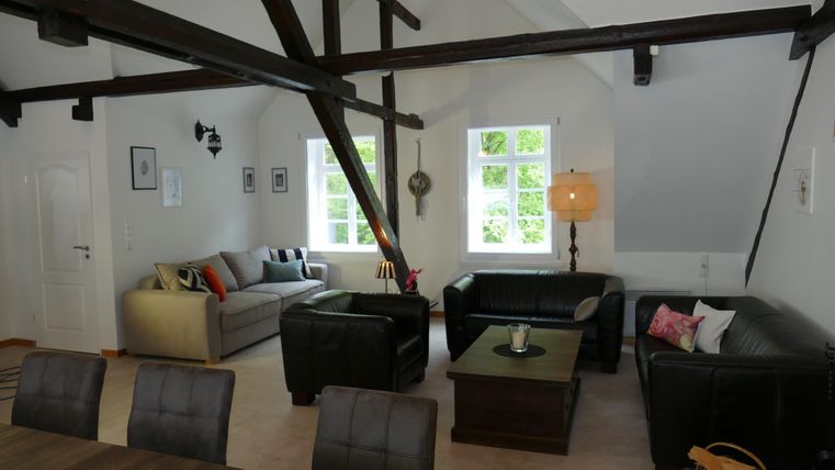 Ein modernes Wohnzimmer mit hohen Decken und sichtbaren Balken. Es gibt gemütliche Sofas, einen Tisch und große Fenster, die viel Licht hereinlassen.