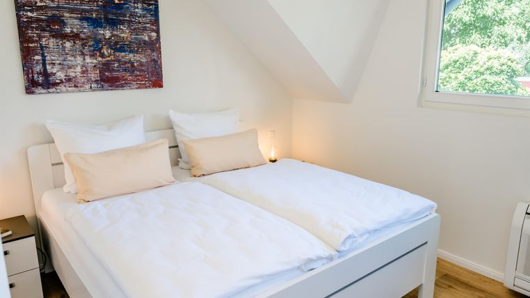 Ein helles Schlafzimmer mit einem Doppelbett und weißen Bettdecken. An der Wand hängt ein modernes Kunstwerk, und das Fenster bietet einen Blick auf die grüne Umgebung.