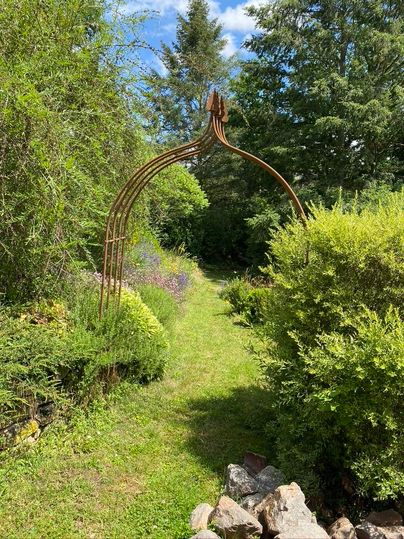 Ein geschmackvoller Gartenweg mit einem rustikalen Tor aus Metall. Umgeben von üppigem Grün und bunten Blumen führt der Weg in die Natur.