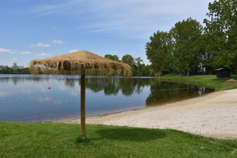Ein ruhiger See mit einem schattenspendenden Strohdach und einer Sandbucht. Im Hintergrund sind grüne Bäume zu sehen.
