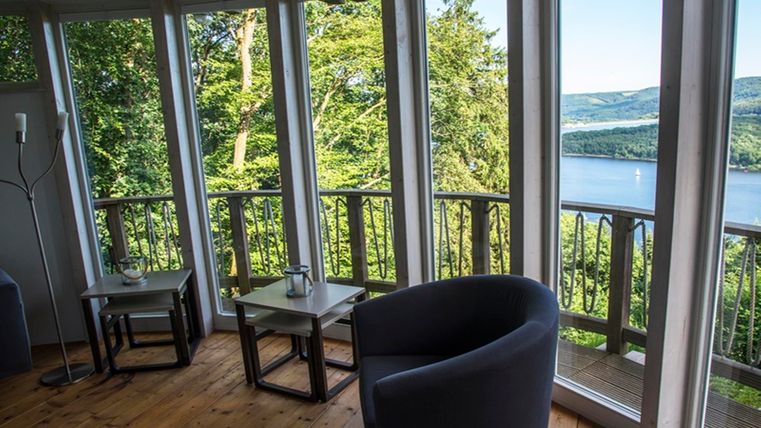 Ein gemütlicher Raum mit großen Fenstern bietet einen Blick auf einen See und die umgebende Natur. Der Holzfußboden und die modernen Möbel schaffen eine einladende Atmosphäre.