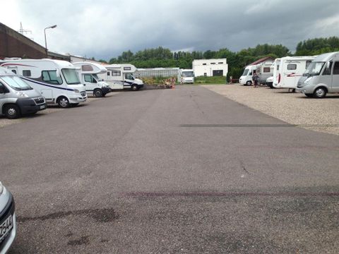 Ein Parkplatz mit mehreren Wohnmobilen und einem grauen Asphaltboden. Im Hintergrund sind Bäume und ein Gebäude zu sehen, während der Himmel bewölkt ist.