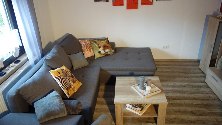 Ein modernes Wohnzimmer mit einer grauen Couch und bunten Kissen. Ein kleiner Holztisch steht in der Mitte, umgeben von einem ansprechenden Bodenbelag.