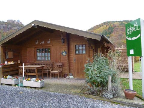 Ein gemütliches Holzhaus mit einer Terrasse und blühenden Pflanzen. Die Umgebung ist von sanften Hügeln und herbstlichen Farben geprägt.