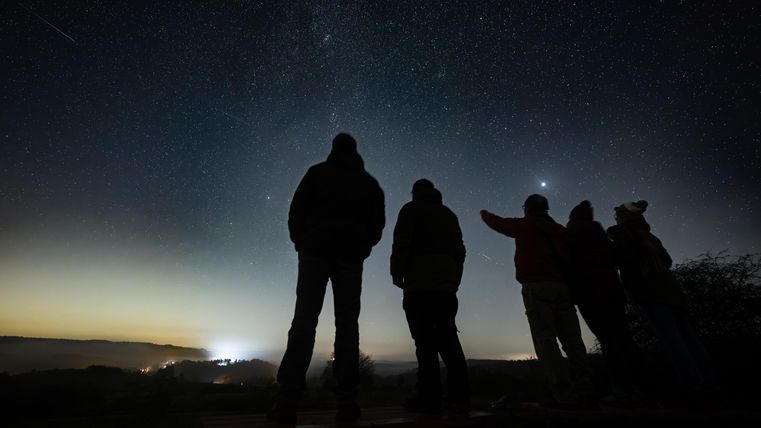 Eine Gruppe von Personen steht im Dunkeln und beobachtet den Sternenhimmel. Der Himmel ist klar und mit vielen Sternen sowie einem leuchtenden Punkt am Horizont.