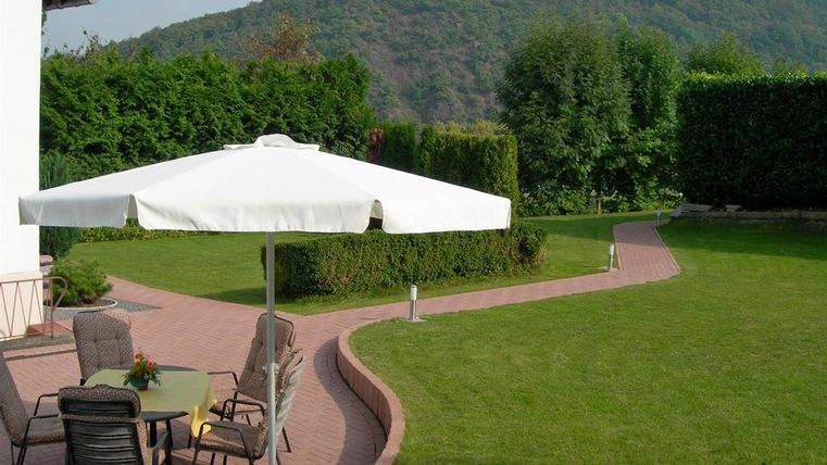 Ein gepflegter Garten mit einer hellen Terrasse und einem großen Sonnenschirm. Im Hintergrund sind grüne Hecken und sanfte Hügel zu sehen.