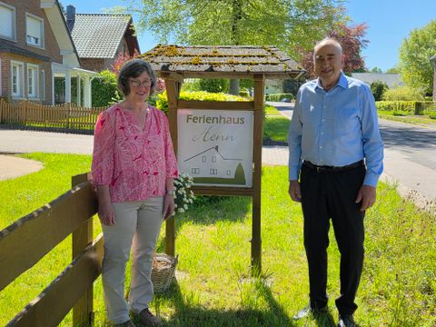 Ein Paar steht neben einem Schild mit der Aufschrift „Ferienhaus eben“ in einer idyllischen Straße. Im Hintergrund sind grüne Bäume und typisch norddeutsche Häuser zu sehen.