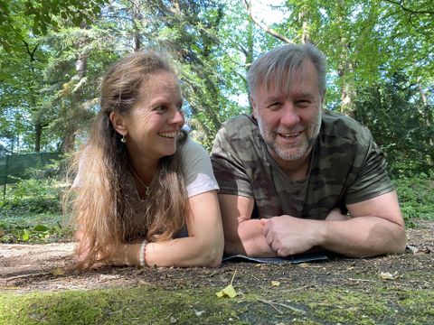Ein Paar liegt entspannt auf dem Boden im Wald und lächelt. Die Umgebung ist grün und von Bäumen umgeben.