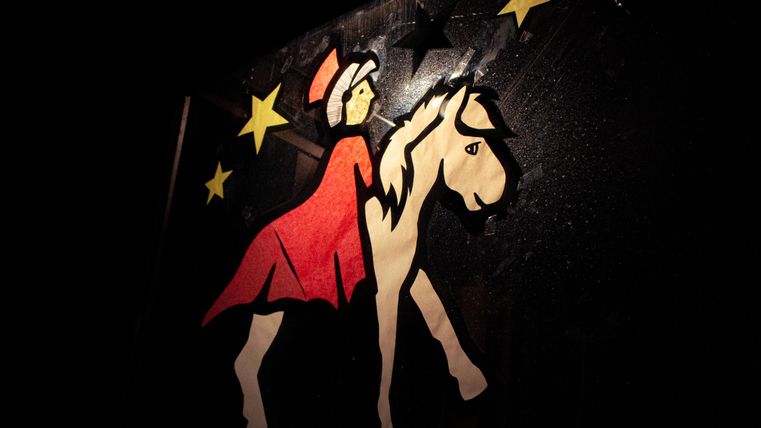 Eine bunte Papierfigur einer Reiterin in einem roten Kleid, die ein Pferd führt. Hintergrund ist dunkel mit goldenen Sternen.