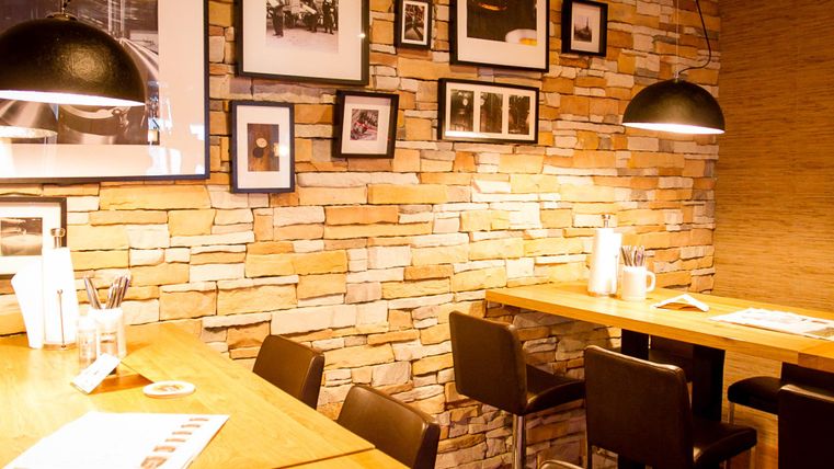 Ein gemütliches Café mit Holztischen und einer Steinfassade. An den Wänden hängen Schwarz-Weiß-Fotos und die Beleuchtung ist warm und einladend.