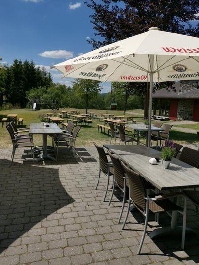 Ein Außenbereich mit Tischen und Stühlen, umgeben von einer grünen Landschaft. Sonnenschirme bieten Schatten an einem sonnigen Tag.