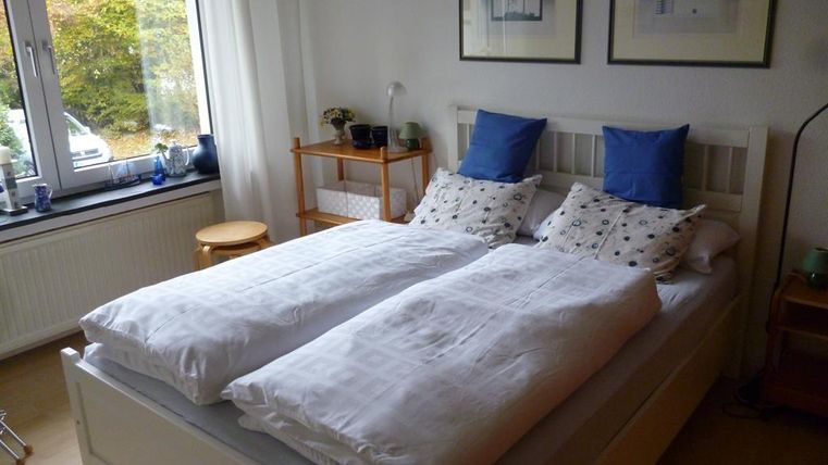 Ein gemütliches Schlafzimmer mit einem weißen Bett und blauen Kissen. Große Fenster lassen viel Tageslicht herein.