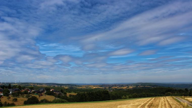 Eine weite Landschaft mit Feldern und sanften Hügeln. Der Himmel ist blau mit einigen Wolken.