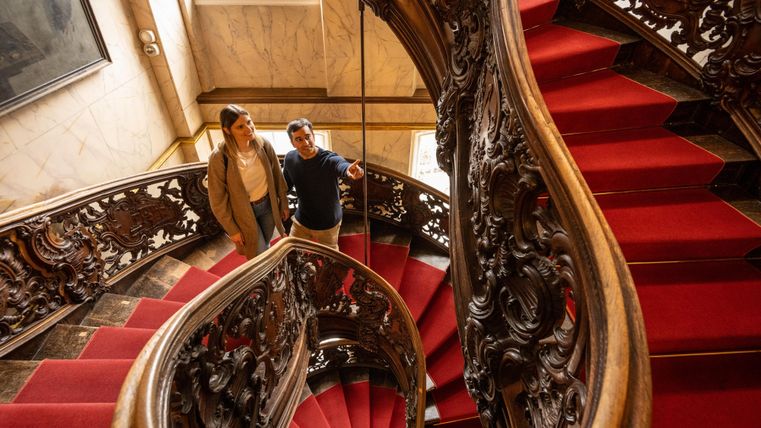 Eine elegante Treppe mit rot gefärbtem Teppich und kunstvollem Holzgeländer. Zwei Personen stehen und schauen nach oben.