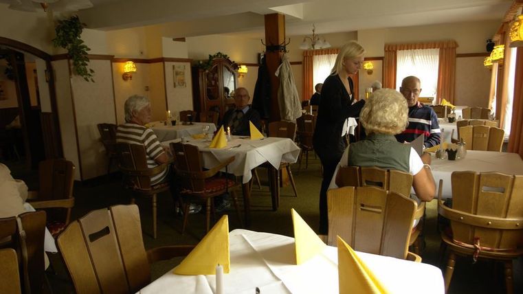 Ein gemütliches Restaurant mit Tischen, die mit weißen Tischdecken und gelben Servietten gedeckt sind. Gäste sitzen und unterhalten sich, während eine Bedienung ihnen die Bestellung aufnimmt.