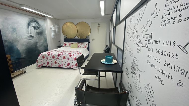 Ein modernes Zimmer mit einem Bett und einer dekorativen Wandgestaltung. Es gibt einen Tisch mit einem Getränk und eine kreative Wand mit handschriftlichen Notizen.