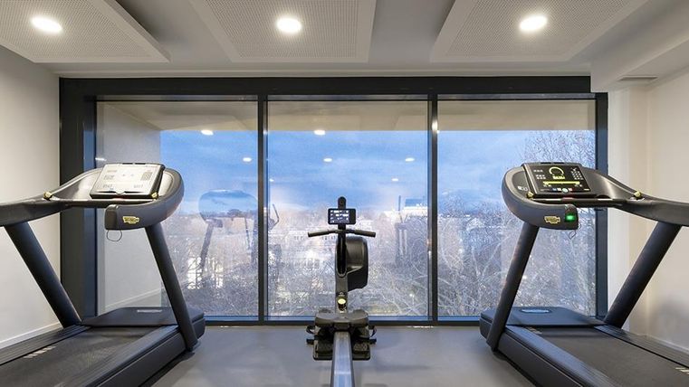 Ein modernes Fitnessstudio mit zwei Laufbändern und einem Fahrradergometer. Große Fenster bieten einen Blick auf die Stadt und den Himmel.