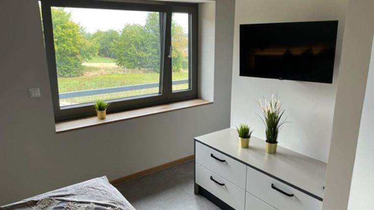 Ein modernes Zimmer mit einem großen Fenster, das auf eine grüne Landschaft blickt. An der Wand hängt ein Fernseher, und auf dem Möbelstück stehen zwei kleine Pflanzen.