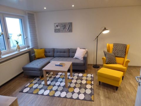 Ein modernes Wohnzimmer mit einer grauen Couch und einem gelben Sessel. Der Raum ist hell und freundlich gestaltet, mit einem Tisch und einem bunten Teppich.