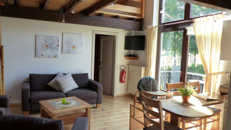 Ein helles Wohnzimmer mit einer bequemen Couch und einem Esstisch. Große Fenster bieten einen Blick auf die grüne Umgebung.