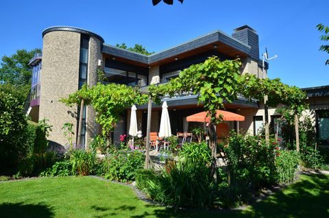 Ein modernes Haus mit großen Fenstern und einem grünen Garten. Im Vordergrund stehen Sonnenliegen und Sonnenschirme unter Weinreben.